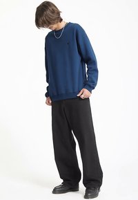 Felpa blu navy con collo a girocollo, polsini a costine e un piccolo logo sul petto, abbinata a pantaloni neri larghi e scarpe nere.