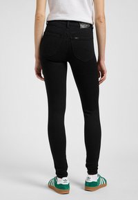 Črne skinny jeans hlače z visokim pasom, ki imajo klasičen dizajn s petimi žepi in subtilnim šivom, v kombinaciji z zelenimi semiš čevlji.