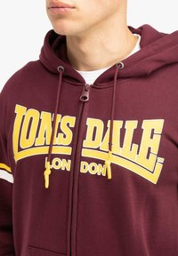 Burgundská mikina na zips so žltým logom "LONSDALE LONDON" a šnúrkami. Hrubá tkanina s hladkým povrchom a klokania kapsa.