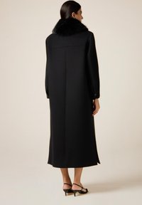 Cappotto lungo nero con colletto in pelliccia nera morbida, design su misura e polsini con bottoni, presentato in uno stile minimalista su uno sfondo neutro.