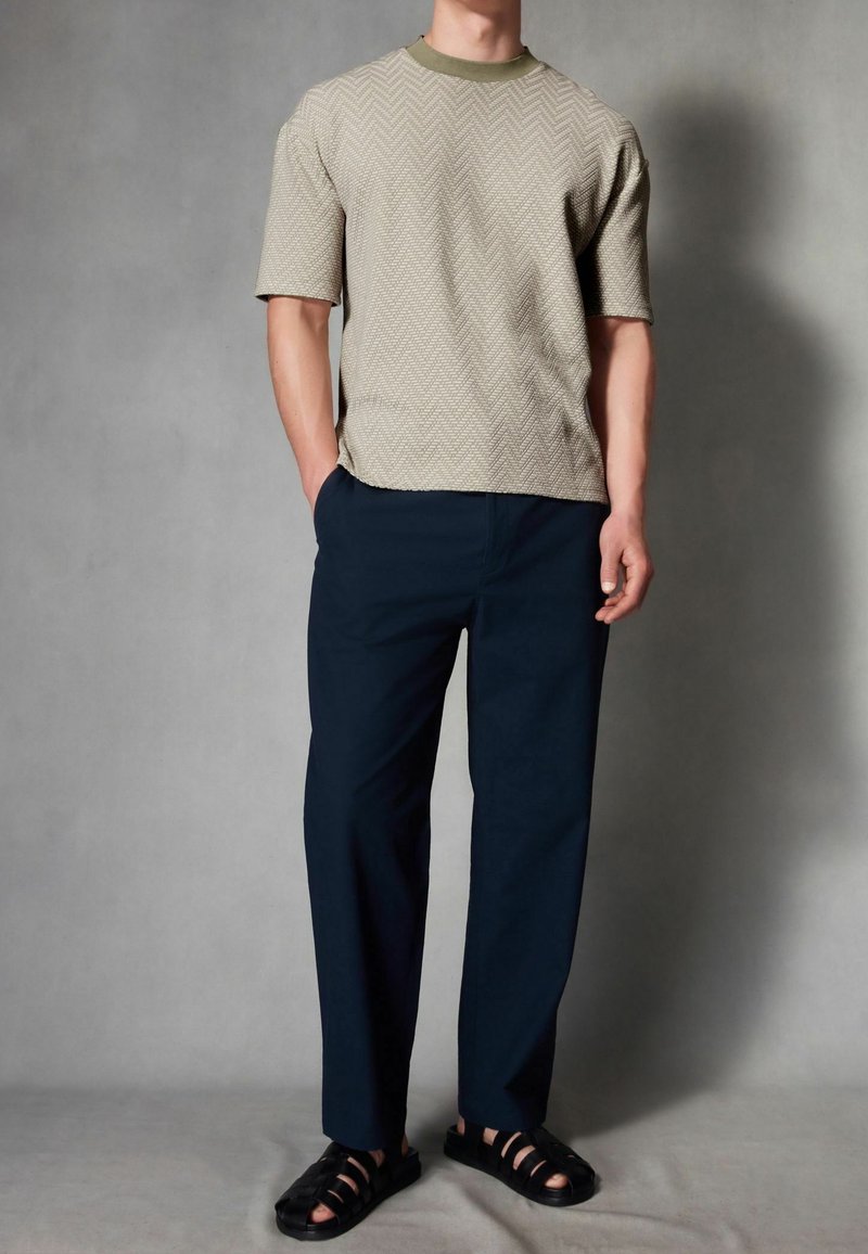 Homme portant une chemise texturée beige à manches courtes, pantalon bleu marine et sandales noires tissées, debout devant un fond gris uni.