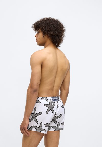 Homme avec des cheveux bouclés portant un short de bain blanc à motif de grandes étoiles de mer noires, se tenant de profil contre un fond clair uni.