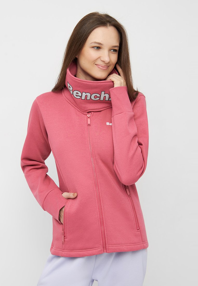 Roze zip-up jack met hoge kraag en "Bench" logo. Beschikt over twee zijzakken en een soepele stoftextuur. Voorzien van een ritsluiting aan de voorkant.