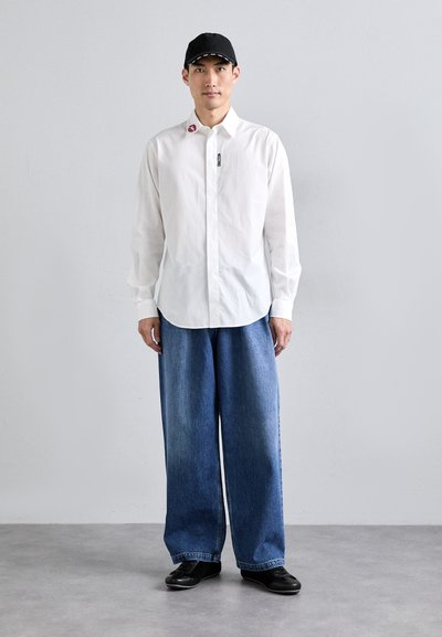 White long-sleeve shirt with a collar and logo, paired with wide blue denim pants and black shoes. Casual cap completes the outfit.