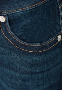 Donkerblauwe spijkerbroek met een voorzak voorzien van contrasterende oranje stiksels en een zilveren metalen klinknagel als accent. Gladde textuur.