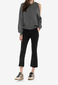 Młoda kobieta ubrana w szary sweter z odkrytymi ramionami, czarne spodnie 3/4 i beżowe trampki z czarnymi sznurówkami, stoi z jedną ręką w kieszeni.