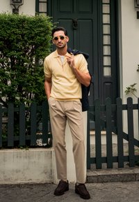 Polo-shirt zippé jaune, pantalon beige, chaussures marron foncé, avec une veste bleu marine drapée sur une épaule. Arrière-plan de haie verte et de porte sombre.