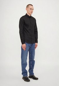 Camisa preta de manga comprida com botões, de textura suave, combinada com jeans de ganga azul. O modelo calça botas pretas de couro. Design simples.