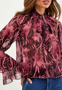 Läpinäkyvä mörkblå blus med rosa blommönster, långa utsvängda ärmar och en rosett knuten runt halsen, i kombination med blåjeans.