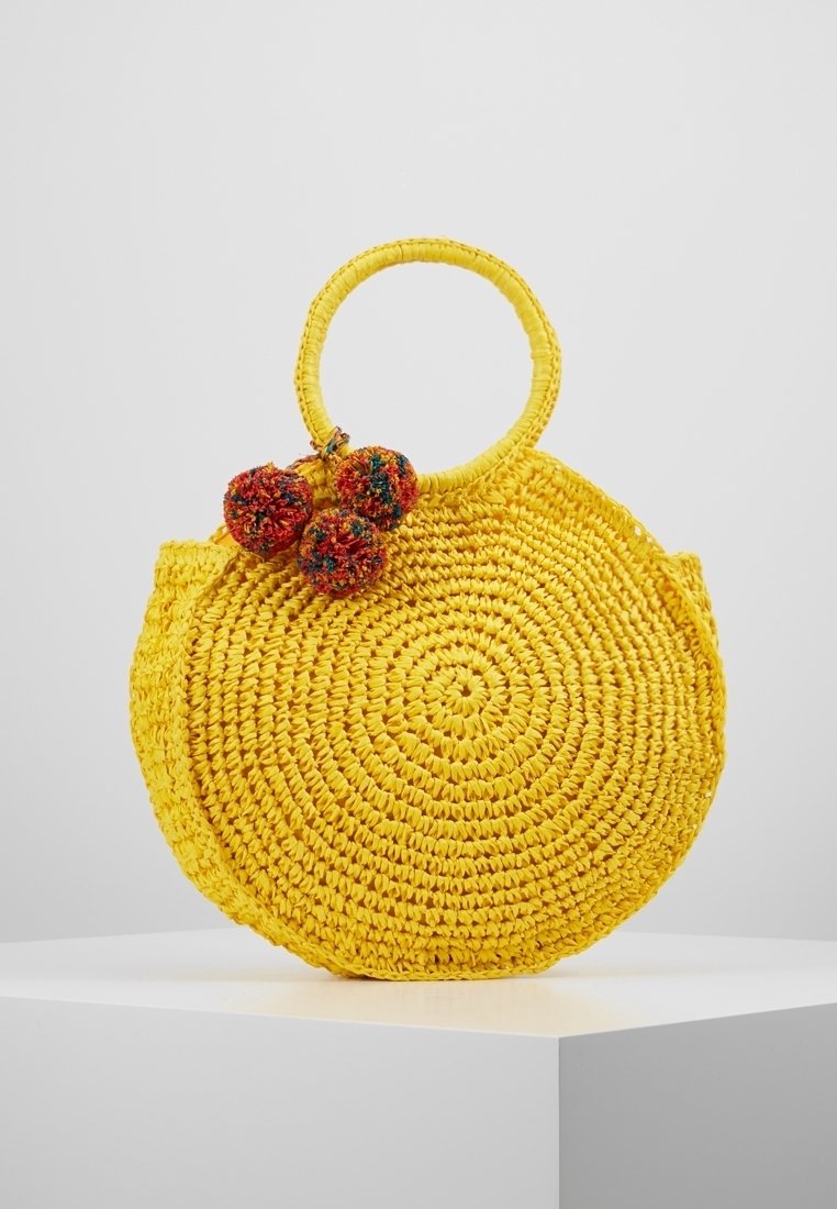Okrągła żółta torba ze słomy z teksturowanym, plecionym wzorem, z dwiema kolorowymi pomponami przymocowanymi do uchwytu.