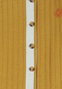 Mosterdkleurige stof met verticale stiksels heeft een witte knoopsluiting en grote ronde knopen. Een geborduurd zonnestraal-detail is zichtbaar.