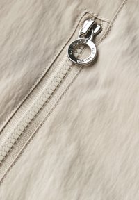 Beige tyg med en slät textur har en silverdragkedja med en rund dragknapp och off-white tänder, vilket visar upp fina sömnadsdetaljer.
