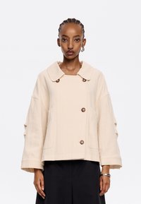 Giacca oversize beige con tessuto testurizzato, grande colletto e due bottoni marroni sul davanti. Presenta tasche laterali e maniche larghe.