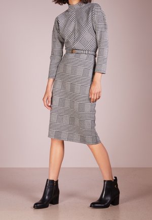 Femme portant une robe à carreaux noir et blanc ceinturée, manches longues, longueur genoux, avec des bottines noires, debout sur un sol uni.