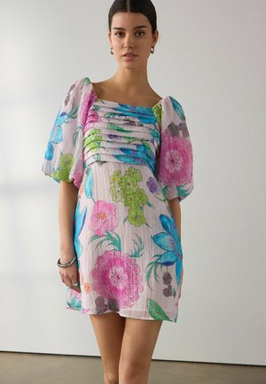 Frau in einem kurzen Kleid mit Puffärmeln und einem großen Blumenmuster in Pink, Blau und Grün, die vor einer schlichten hellen Wand steht.