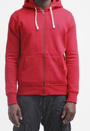 Mann trägt einen roten Kapuzenpullover mit Reißverschluss und weißen Kordeln sowie schwarze Cargohosen und steht vor einem weißen Hintergrund.