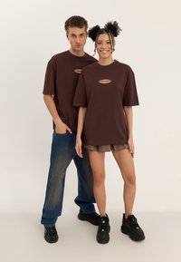 T-shirts en coton marron avec un logo au centre, design oversize. Associés à un jean bleu et une mini-jupe à carreaux. Les deux portent des baskets noires.