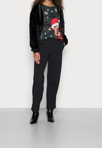 Veste bomber en velours noir avec des détails en satin, pull de Noël vert avec un motif de renne, et pantalon noir ajusté à coupe droite.