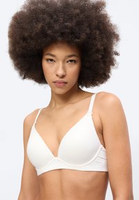 Νεαρή γυναίκα με μεγάλα σγουρά αφρό μαλλιά που φοράει λευκό σουτιέν, κοιτάζοντας απευθείας την κάμερα πάνω σε απλό ανοιχτό φόντο.