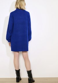 Asmuo vilkintis karališkai mėlyną megztinį su suknele ir juodomis vidutinio ilgio kulninėmis botais, stovintis prieš paprastą baltą sieną.