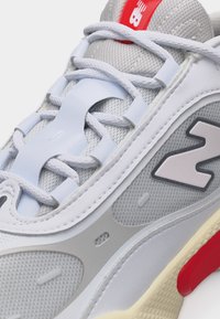 Λευκό και γκρι αθλητικό παπούτσι με κόκκινες λεπτομέρειες, λευκά κορδόνια, ύφασμα από δίχτυ και μεγάλο λογότυπο "N" στην πλευρά.