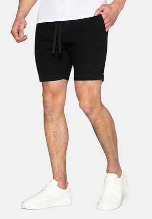 Homme portant un short noir à cordon et des baskets blanches, debout avec une main dans la poche sur un fond uni.