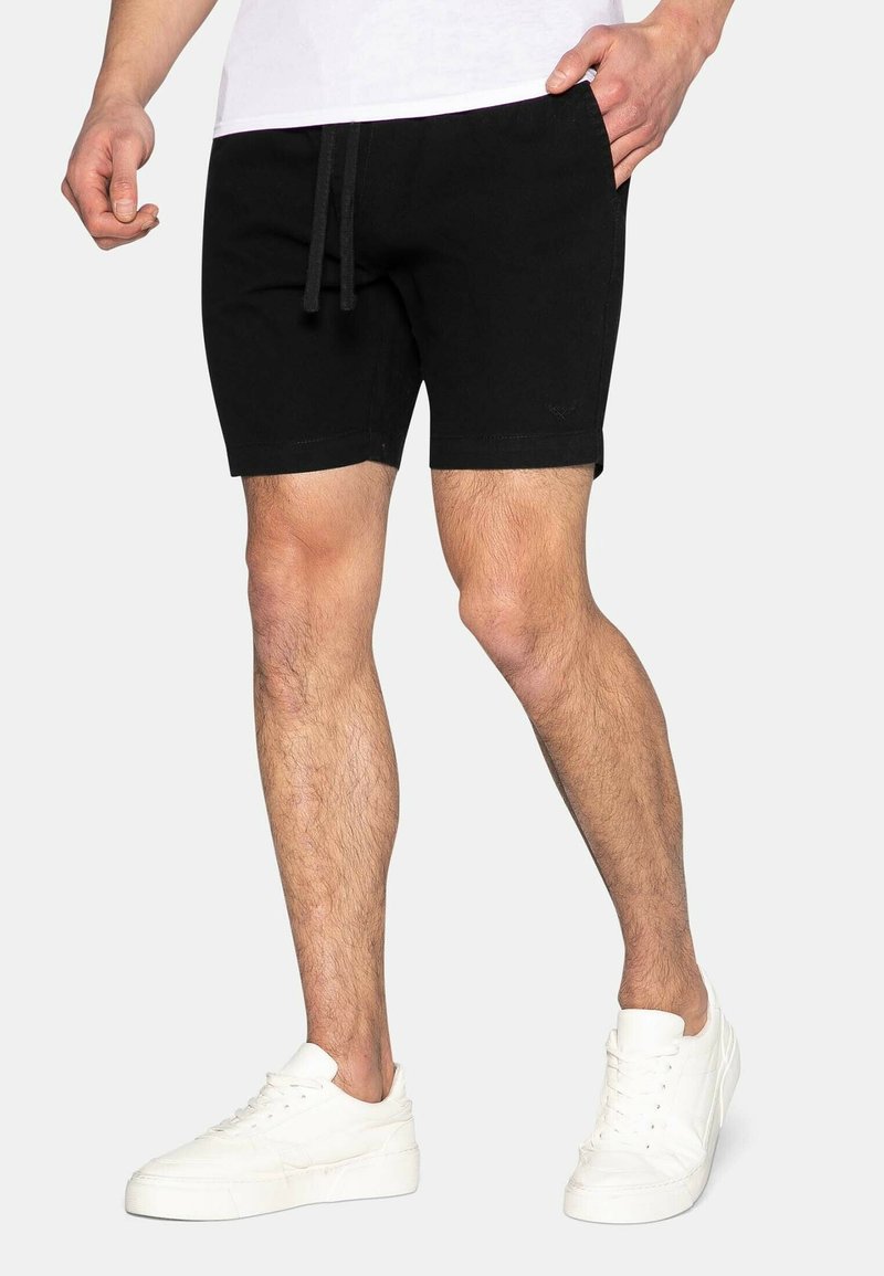 Homme portant un short noir à cordon et des baskets blanches, debout avec une main dans la poche sur un fond uni.