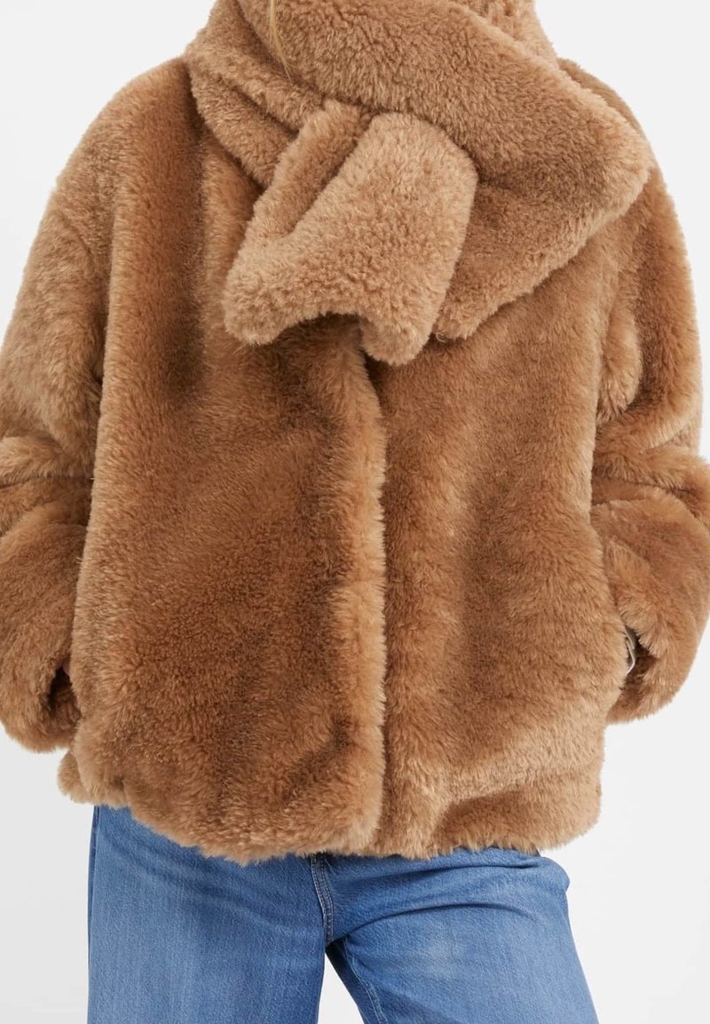 Personne portant une veste en fausse fourrure marron pelucheuse et une écharpe assortie, associées à un jean en denim bleu sur fond blanc.