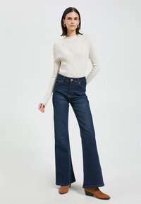Jasnobeżowy sweter prążkowany z teksturowanym wzorem w zestawie z ciemnoniebieskimi dżinsami dzwonami. Model nosi brązowe botki za kostkę. Prosty, elegancki design.
