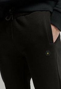 Zwarte sweatpants van zachte stof, met een verstelbare tailleband en een hexagonale patch met gele accenten op de zak.