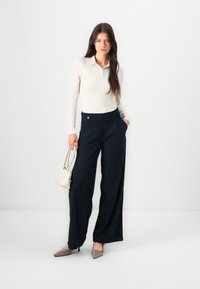 Pantaloni ampi blu navy con una finitura testurizzata, vita alta e dettaglio dei bottoni; abbinati a un polo attillato a maniche lunghe color crema e una borsa leggera.