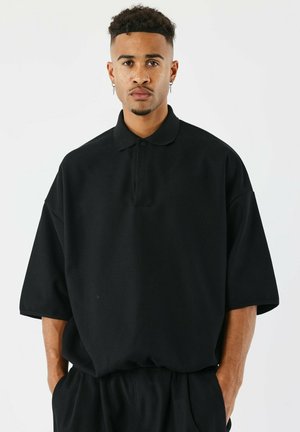 Polo shirt nera oversize con maniche corte, dotata di colletto con bottoni e tessuto texture. Abbinato a pantaloni neri dalla vestibilità rilassata.