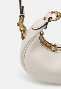 Gros plan d’un sac à main en cuir blanc avec des éléments en laiton, comprenant une fermeture éclair, un fermoir et des détails de chaîne sur la fixation de la bandoulière.