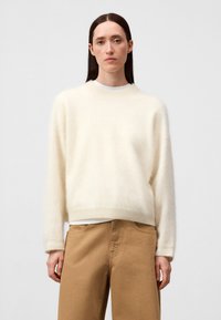 Weicher Cremepullover mit lässiger Passform, rundem Hals und langen Ärmeln, über einem weißen Hemd getragen. Das Material wirkt fusselig und hat eine glatte Textur.