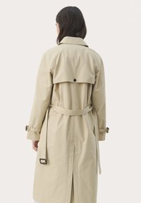 Bež trench coat, mille tagaklapiga, seotud vöö ja rihmade aktsentidega varrukatel. Sile kangas klassikalise ja eritellimusel valitud disainiga.