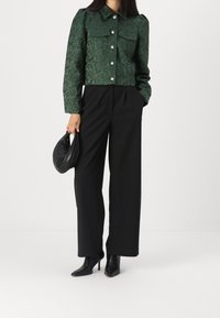 Grön kort jacka med blommig textur, silverknappar och två bröstfickor, parat med högmidjade svarta byxor och svarta klackstövlar.