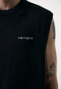Črna brezrokavna majica z rebrastim ovratnikom, na prsih ima belo "Carhartt" oznako, gladka tekstura tkanine, vidna tetovaža na roki.