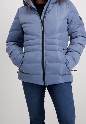 Vrouw draagt een lichtblauwe doorgestikte pufferjas met ritssluiting aan de voorkant en handen in de zakken, gecombineerd met donkerblauwe jeans.