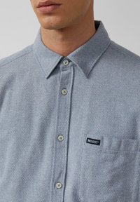 Camicetta azzurra chiara con motivo testurizzato, due bottoni, una tasca con etichetta logo e un design di colletto classico.
