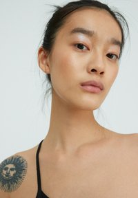 Zwarte tanktop, gladde huid, natuurlijke make-up, prominente zonnetattoo op de schouder, minimaal haar naar achteren vastgezet, neutrale achtergrond.