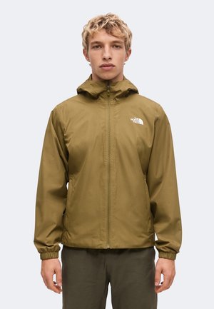 Jeune personne aux cheveux blonds courts et bouclés portant une veste à capuche vert olive avec le logo The North Face sur la poitrine.