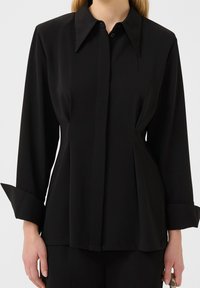 Chemise noire à boutons avec manches longues, grand col et taille ajustée. Tissu lisse avec détails plissés et larges poignets.