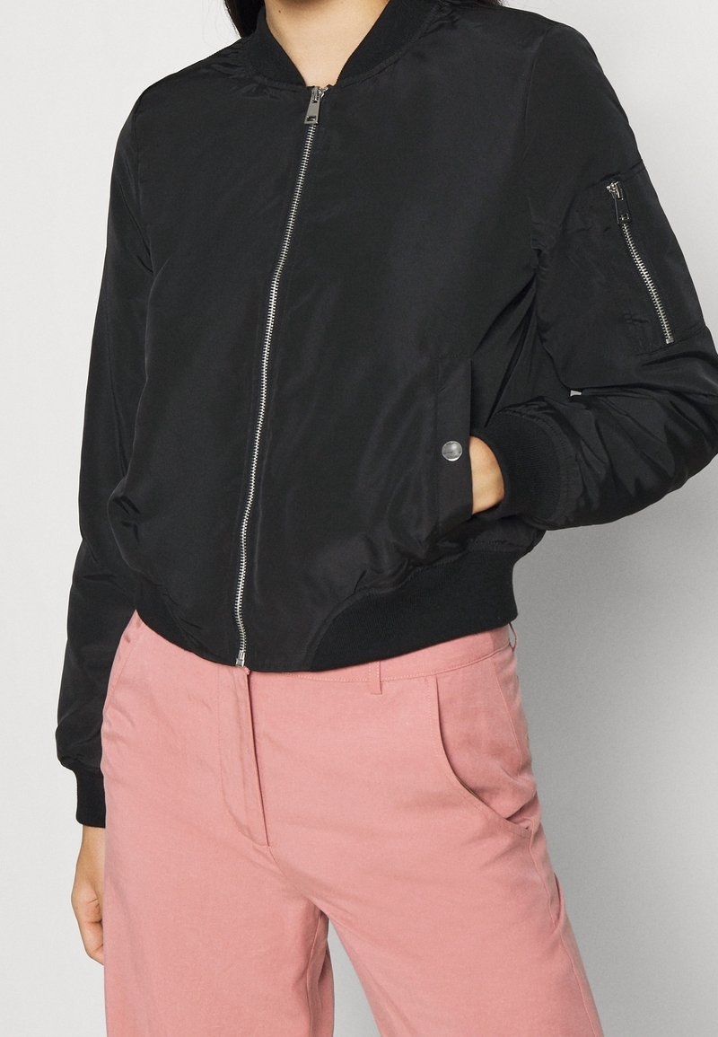 Veste bomber noire avec fermeture éclair à l'avant, poignets côtelés et poches latérales. Comprend une poche à manches zippée et une texture de tissu lisse et mate.