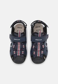 Zapatos sintéticos azul y negro con malla, con bandas de Velcro, acentos a rayas rojas y blancas, lados abiertos y punteras reforzadas.