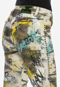 Jean éclaboussé de peinture jaune, turquoise et noire, avec des clous métalliques sur le côté et des lettres en style graffiti au dos et sur la poche de la jambe.