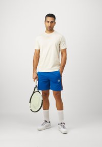Meeste spordirõivad, milles on kreemivärvi ümar kaelusega T-särk ja sinised lühikesed püksid külgribaga, hoides tennisereketit, valgeid tennisetossusid ja sokke.