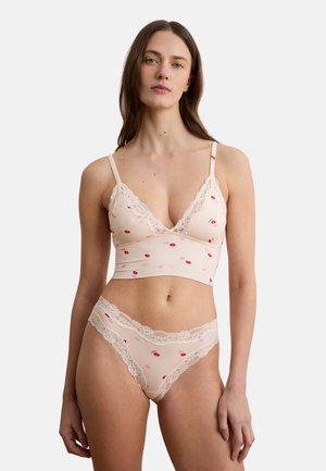 Frau trägt hellrosa Spitzen-Bralette und passende hochgeschnittene Panty mit rotem und pinkem Lippenmuster auf weißem Hintergrund.
