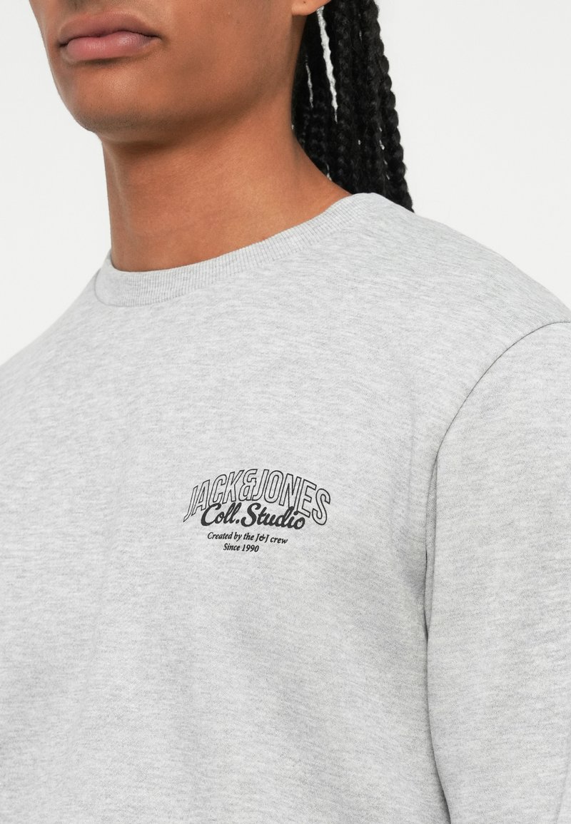 Sudadera gris de material suave, con un cuello redondo y un estampado de logotipo negro en el lado izquierdo del pecho, acompañado de acentos de texto en curva.