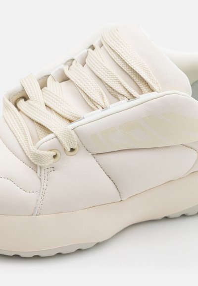 Beige sneaker with a smooth leather upper, textured laces, and subtle logo detailing. Thick rubber sole and rounded shape.