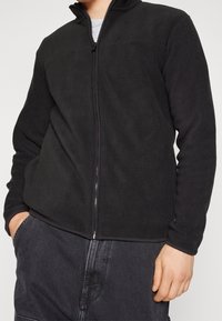 Chaqueta de forro polar negra con cuello alto, cierre de cremallera, mangas largas y textura suave. Se lleva con pantalones grises que tienen bolsillos.